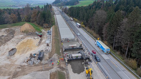 Blick auf die Baustelle der Autobahn A1 im Streckenabschnitt Mondsee–Thalga. Man sieht einen Gleitschalungsfertiger beim Einbau einer Betondecke, Arbeiter überwachen diesen Vorgang. Links davon sieht man gelagerte Baumaterialien, rechts davon sieht man den laufenden Autoverkehr.